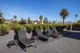 Located at 710 N El Centro Ave in the heart of Hollywood, Los Angeles, this contemporary rooftop amenity space offers an exceptional outdoor experience defined by clean architectural lines and resort-style features. The rooftop is thoughtfully designed with a combination of composite wood decking and large-format tile, creating distinct zones for relaxation and recreation. A sparkling pool serves as the centerpiece, accompanied by a spa, and surrounded by sleek lounge chairs positioned to take full advantage of the open California sky and sweeping panoramic views of the surrounding Hollywood neighborhood, including palm tree-dotted skylines and distant hillsides. Glass panel railings line the perimeter, maintaining unobstructed sightlines while ensuring safety. Generously sized planter boxes filled with drought-tolerant and tropical plantings, including dramatic palm trees and colorful succulents, add vibrant greenery and a sense of privacy throughout the space. A modern elevator housing structure provides direct rooftop access, finished in smooth stucco consistent with the building's contemporary aesthetic. The courtyard level, visible in the 3D overview, reveals additional landscaped outdoor gathering areas shaded by mature palms, further enhancing the property's resort-like atmosphere. Abundant natural light and open-air exposure make this rooftop a standout communal feature of the property.