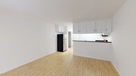 This single-floor unit features a clean, minimalist design throughout. The open living area is finished with light wood-tone flooring and crisp white walls, creating a bright and airy atmosphere. A large sliding glass door fitted with vertical blinds fills the space with natural light and provides a view of the outdoors. A wall-mounted air conditioning unit serves the living area.

The kitchen is thoughtfully designed with white shaker-style cabinetry offering generous upper and lower storage. Dark granite countertops contrast the white cabinetry, and a peninsula with a matching dark countertop separates the kitchen from the living area. Stainless steel appliances include an electric range, an over-the-range microwave, a refrigerator, and a double basin sink. White tile flooring runs through the kitchen area.

The bathroom is well-appointed with a dark granite vanity countertop, an undermount sink with chrome fixtures, a large mirror with a storage cabinet, and a tub and shower combination enclosed by sliding glass doors with chrome hardware. White tile flooring completes the clean, cohesive look of the bathroom.