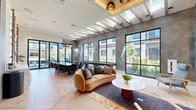 Located at 112 Bilby Road in Hackettstown, NJ, this elegantly designed amenity lounge offers a refined communal experience within a single, open-plan floor. Upon entry through sleek black-framed glass partition doors, guests are greeted by striking light oak herringbone-patterned flooring that flows seamlessly throughout the space. Soaring ceilings with exposed wood beam accents and a row of skylights flood the interior with abundant natural light, creating an airy, upscale atmosphere.

The lounge is anchored by a curved, camel-toned sofa and curated seating arrangement centered around a modern fireplace with a wall-mounted television, ideal for relaxed gatherings. Textured geometric wall paneling adds visual depth, while floor-to-ceiling windows frame lush outdoor courtyard views, further enhancing the connection to the exterior.

A dedicated dining area features a large dark marble-topped table surrounded by leather chairs, illuminated by a sculptural globe pendant chandelier. Alongside it, a shuffleboard table adds a playful social element. A built-in wet bar with glass shelving displays an artful wine and bottle collection, accompanied by a beverage refrigerator and counter space, completing this multifunctional entertaining environment. Statement pendant lighting and curated artwork throughout elevate the space's contemporary, hospitality-inspired design.