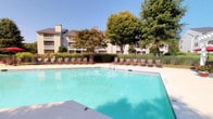 This outdoor amenity space centers around a generously sized swimming pool with clear, blue-green water, offering a refreshing retreat for residents. The pool deck is finished in smooth concrete and lined with rows of cushioned lounge chairs, providing ample space for sunbathing and relaxation. A striking white pergola anchors one end of the deck, offering shaded seating beneath its open lattice roof, complemented by red patio umbrellas and additional outdoor furniture for casual gathering.

The area is enclosed by a wrought iron fence, ensuring a sense of privacy and security, while mature trees and ornamental grasses create a lush, natural backdrop that softens the surroundings. Carefully maintained landscaping, including clusters of tall ornamental grass and flowering shrubs, adds texture and visual interest throughout the space. Well-kept lawns border the pool enclosure, contributing to the overall manicured aesthetic.

The multi-story residential buildings visible in the background, featuring neutral siding, pitched rooflines, and private balconies, frame the space cohesively. Under open, sunny skies, the entire area benefits from abundant natural light, enhancing the inviting and resort-like atmosphere of this communal outdoor space.