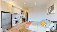 This thoughtfully designed studio presents a seamless open-concept layout where living, sleeping, and cooking areas flow naturally into one another. Light hardwood flooring runs throughout, lending warmth and continuity to the space. Large floor-to-ceiling sliding glass doors open to a private balcony, flooding the interior with natural light and offering open city views, making the studio feel notably airy and spacious.

The kitchen is positioned along one wall and is fitted with light wood cabinetry, white quartz countertops, a white tile backsplash, stainless steel appliances including a full-size refrigerator and built-in microwave, an induction cooktop, and a stainless steel sink with a modern faucet. An in-unit washer and dryer are conveniently integrated into the same wall. Open shelving adds practical display space above the counters.

The main living area comfortably accommodates a full-size bed alongside a seating arrangement near the balcony, where an abundance of natural light enhances the relaxed atmosphere. Track lighting throughout provides flexible, well-distributed illumination.

The bathroom features a clean, minimalist design with a floating white vanity, an undermount sink, a large mirror, and a separate shower area with a curtain enclosure, all finished with consistent light hardwood flooring.