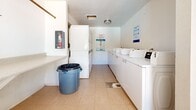 Located at 1022 E Narramore Ave in Buckeye, Arizona, this shared laundry facility serves the residents of The Village at Camelot community. The single-room space is thoughtfully laid out to maximize functionality, with clean white walls and light-toned tile flooring that give the interior a bright, tidy appearance. A row of top-load washing machines lines one wall, while stacked washer-dryer units are positioned along the opposite side, providing ample laundry capacity for the community. A built-in folding counter runs along one wall, offering a convenient surface for sorting and folding laundry. Natural light filters into the space through a window and a glass-paned exterior door, which also provides direct outdoor access. The door features a ventilation panel at its base, supporting airflow throughout the room. A flush-mounted ceiling light ensures consistent illumination. Posted signage includes laundry tips and community use guidelines, reinforcing that the facility is reserved exclusively for tenants. A utility panel is also present, keeping essential building systems accessible. The overall layout is clean, efficient, and well-equipped to meet the day-to-day laundry needs of residents.