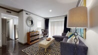 This thoughtfully designed single-floor residence showcases a cohesive modern aesthetic carried throughout each room. The living area serves as the heart of the space, featuring dark hardwood-style flooring, crown molding, and a large sliding door dressed with dramatic floor-to-ceiling black curtains and sheer vertical blinds that allow natural light to filter in. A bold geometric patterned area rug anchors the room, complemented by a plush gray sectional sofa, a tufted ottoman, and an iconic leather egg chair — all contributing to a sophisticated yet comfortable atmosphere.

The bedroom continues the warm, curated design with teal drapery framing a bright window, dark hardwood-style flooring, and a ceiling fan overhead. A wooden dresser with a round mirror adds classic character to the room.

The kitchen is efficiently laid out in a U-shape configuration, offering gray cabinetry, granite-style countertops, and a full suite of stainless steel appliances including a refrigerator, range, and dishwasher. A window above the sink brings in natural light.

The bathroom features an extended gray vanity with a wide granite-style countertop, a large dark-framed mirror, and Hollywood-style vanity lighting. Dark hardwood-style flooring ties the space together, and colorful artwork adds a vibrant finishing touch.