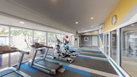 Located at 14901 E Mississippi Ave in Aurora, Colorado, this well-appointed fitness center offers a bright, modern environment designed to support a full range of workout activities. The single-floor space is flooded with natural light through a generous wall of floor-to-ceiling windows along one side, providing views of the surrounding greenery and creating an open, energizing atmosphere. Recessed lighting and a skylight further illuminate the interior throughout the day.

The main exercise area features a lineup of cardio machines, including treadmills and elliptical trainers, positioned to take advantage of the natural light and outdoor views. Fitness balls are available for supplementary training. A dedicated open-floor stretching and flexibility zone is finished with smooth flooring, complemented by a bold geometric mural in shades of blue, teal, yellow, and white that adds a dynamic visual element to the space.

The striped, multi-tone carpet in the main workout zone brings a contemporary design touch, while the neutral gray walls provide a clean, professional backdrop. A wall-mounted television is available for entertainment during workouts. Fresh towels are conveniently stocked on a shelving unit near the entrance, and the space is equipped with a wall-mounted air conditioning unit for climate control. Direct exterior access is provided through a glass door, connecting the fitness center seamlessly to the outdoor courtyard area.