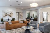 This single-floor amenity lounge is designed to serve as a versatile communal gathering space, blending comfort and functionality through thoughtful design. The entire area is unified by striking herringbone-patterned hardwood flooring, which runs continuously throughout every zone and contributes warmth and visual cohesion to the open layout.

The central lounge area serves as the focal point of the space, anchored by a textured tile fireplace framed with decorative artwork. A mix of seating — including rounded swivel chairs in heathered grey, a cognac leather sofa, and mid-century accent chairs — surrounds a marble-topped coffee table, creating an inviting conversation area beneath a large drum pendant light.

Adjacent to the lounge, a casual dining and seating zone features bistro-style round tables paired with upholstered chairs, illuminated by matte black pendant lights. Large windows throughout admit ample natural light and offer views of the surrounding greenery.

A well-appointed wet bar station includes charcoal shaker-style cabinetry with brushed gold hardware, a white quartz countertop, undermount sink, stainless steel appliances, and open shelving backed by a light grey subway tile. A full-size stainless steel refrigerator is also available nearby.

A dedicated game area houses a foosball table, completing this multi-functional space designed for both relaxation and recreation.