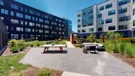 This outdoor amenity courtyard is nestled within a contemporary multi-story residential complex, defined by striking dark brick and light panel façades that frame the space on multiple sides. The courtyard is thoughtfully designed to offer a variety of recreational and relaxation zones across its expansive paved and landscaped grounds.

At the heart of the space, a concrete-top ping-pong table with a bold wooden base serves as a central recreational feature, positioned along a wide, gray stone-paved pathway that guides residents through the area. Adjacent to it, a seating arrangement with plush outdoor sofas, accent stools, and a fire pit provides an inviting lounge area ideal for socializing.

Further into the courtyard, a shaded dining zone with navy blue shade sails covers a collection of tables and chairs, offering a comfortable outdoor dining experience. A well-maintained lawn area is outfitted with lounge chairs, perfect for relaxing under open skies. Lush landscaping throughout—including flowering shrubs, ornamental grasses, young trees, and decorative rock beds—adds a layer of natural beauty and texture to the space.

Large glass entry doors connect the interior building to the courtyard, ensuring seamless indoor-outdoor flow. The overall ambiance is modern, vibrant, and community-oriented, designed to encourage outdoor living and social engagement among residents.