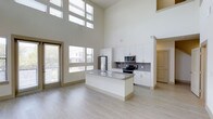 This two-story Dallas property showcases a dramatic open-concept layout defined by soaring double-height ceilings that create an expansive, airy atmosphere. The main living area is flooded with natural light through a generous arrangement of large windows and a glass door, offering views of the surrounding neighborhood while maintaining a bright, welcoming ambiance throughout the day.

The kitchen serves as the centerpiece of the main floor, thoughtfully designed with crisp white cabinetry extending to considerable height along the walls. A large central island with a granite countertop provides ample workspace and casual seating potential, while stainless steel appliances — including a French door refrigerator, electric range, and built-in microwave — deliver a sleek, contemporary finish. A gray tile backsplash adds a subtle textural contrast to the clean white cabinetry.

Light wood-toned flooring runs throughout the main level, complementing the neutral color palette and enhancing the sense of openness. A staircase visible toward the rear of the space leads to the second floor. The overall design aesthetic is modern and minimalist, prioritizing clean lines, natural light, and functional layout.