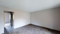 This single-floor space presents a straightforward and functional layout designed with simplicity and practicality in mind. The interior is finished with dark hardwood-style plank flooring that runs throughout, providing a warm contrast against the light, neutral-toned walls. The textured popcorn ceiling adds a classic touch overhead, while crisp white baseboards frame the perimeter of each room, contributing to a clean and cohesive aesthetic. The walls are painted in a soft, understated gray-white tone that creates a bright and open atmosphere, allowing for versatile interior design possibilities. The space is well-proportioned, offering a comfortable sense of openness without feeling overwhelming. A doorway visible in the corner suggests a connected, flowing floor plan that links the main living areas efficiently. The overall ambiance is clean, neutral, and move-in ready, serving as a blank canvas for a variety of furnishing styles and personal touches. The minimalist finish palette ensures that the space can easily adapt to both modern and traditional décor preferences, making it a highly versatile and practical living environment.