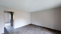 This single-floor space presents a straightforward and functional layout designed with simplicity and practicality in mind. The interior is finished with dark hardwood-style plank flooring that runs throughout, providing a warm contrast against the light, neutral-toned walls. The textured popcorn ceiling adds a classic touch overhead, while crisp white baseboards frame the perimeter of each room, contributing to a clean and cohesive aesthetic. The walls are painted in a soft, understated gray-white tone that creates a bright and open atmosphere, allowing for versatile interior design possibilities. The space is well-proportioned, offering a comfortable sense of openness without feeling overwhelming. A doorway visible in the corner suggests a connected, flowing floor plan that links the main living areas efficiently. The overall ambiance is clean, neutral, and move-in ready, serving as a blank canvas for a variety of furnishing styles and personal touches. The minimalist finish palette ensures that the space can easily adapt to both modern and traditional décor preferences, making it a highly versatile and practical living environment.