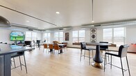 Located at 29 Sixth St NE in Rochester, Minnesota, this community room offers a well-appointed, single-floor open-plan layout that combines a fully equipped kitchen with a spacious living area. Light wide-plank hardwood flooring flows throughout, unifying the space and complementing its contemporary industrial aesthetic, which includes exposed ductwork and track lighting.

The kitchen is outfitted with light wood cabinetry, stainless steel appliances including a double wall oven and a large French door refrigerator, and a dark-toned island with pendant lighting overhead. The adjacent living area features a substantial sectional sofa, a bold stone accent wall with an integrated fireplace and mounted displays, and a striking patterned area rug that anchors the lounge setting.

A separate dining and multi-use gathering zone provides multiple seating configurations, including round tables with mixed seating and bar-height tables with stools, making it well-suited for both casual and organized group events. Large windows lining the perimeter flood the interior with natural light while framing panoramic views of the Rochester skyline. A glass door opens directly onto an outdoor terrace, extending the usable space and providing additional seating with city views. The overall ambiance is modern, inviting, and versatile.