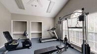 Located in Douglasville, GA, this fitness center offers a dedicated exercise room designed to support a well-rounded workout routine. The space features vaulted ceilings that lend an open, airy feel despite its compact footprint, while a ceiling fan and overhead fluorescent lighting ensure comfort during exercise sessions. Natural light streams in through large windows fitted with blinds, including a distinctive arched window that adds an architectural touch to the room. The walls are finished in a neutral gray tone, complemented by blue-gray carpeted flooring that runs throughout. Built-in open shelving units along one wall provide convenient storage for equipment and personal items. The exercise room is equipped with a variety of fitness machines, including a treadmill, a recumbent stationary bike, an elliptical trainer, and a multi-function cable weight machine with an adjustable bench, catering to both cardiovascular and strength training needs. A large wall-mounted mirror expands the sense of space and allows users to monitor their form. A French door entry adds a refined touch to the overall design, making this fitness center a functional and well-appointed space for daily exercise.