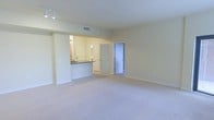 This single-floor space offers a practical and well-organized layout designed to maximize functionality. The living area is finished with plush wall-to-wall carpeting in a neutral tone, contributing to a clean and understated aesthetic throughout. Smooth, light-colored walls and white trim create a bright, airy atmosphere, complemented by recessed ceiling lighting and a track light fixture that provide even, adjustable illumination.

A defining feature of the space is the pass-through kitchen opening, which connects the kitchen to the living area. The kitchen is visible through this opening and features dark countertops that contrast with the lighter cabinetry and white louvered cabinet doors, adding a classic touch to the space. The central HVAC system is served by a ceiling vent, ensuring efficient climate control throughout.

Two interior doorways are visible from the main living area — one leading to an adjacent room and another opening to a hallway, suggesting additional rooms beyond. The overall design is straightforward and move-in ready, with a cohesive neutral palette that offers a versatile backdrop for a variety of interior styles.