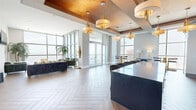 Located at Rocky Point in Tampa, Florida, this sophisticated amenity space spans a generous single floor and is designed to impress at every turn. The layout encompasses two distinct living areas, seamlessly blending interior elegance with outdoor living. The main interior lounge features striking herringbone wood-look flooring, a coffered ceiling with warm gold leaf accents, and a series of pendant chandeliers with brass detailing. A bold geometric-tiled accent wall frames a built-in bar with dark cabinetry, open shelving, and green velvet bar seating, adding a vibrant yet refined focal point to the room. Floor-to-ceiling glass walls flood the space with natural light and frame panoramic views of the surrounding waterway and Tampa skyline. A second living area opens directly onto a covered outdoor terrace finished in large-format stone tile, where a patterned tile fireplace wall and lounge seating create a comfortable alfresco retreat. Additional open-air terrace space offers dining areas and sweeping, unobstructed views of the water and horizon. Throughout, the design balances modern architecture with luxurious finishes, delivering a dynamic and visually captivating environment ideal for gathering and relaxation.