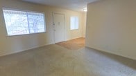 This single-floor residential space offers a clean, functional layout designed for comfortable everyday living. The main living area is a defining feature of the home, showcasing a combination of soft, neutral-toned carpeting and hardwood-style flooring that transitions naturally between zones, creating a sense of warmth and definition throughout the open floor plan. The walls are finished in a light, creamy tone that enhances the brightness of the space and complements the natural light streaming in through multiple windows fitted with vertical blinds. These windows provide both privacy and the ability to control light levels throughout the day. The front entry door opens directly into the living area, creating an accessible and welcoming flow from the moment you step inside. The overall ambiance is bright, neutral, and move-in ready, offering a versatile canvas that can be adapted to a wide range of interior styles and furnishing preferences. The simplicity of the design allows the natural light and open layout to take center stage, making the space feel airy and uncluttered. With its straightforward single-floor layout and practical finishes, this space is well-suited for those seeking a low-maintenance, comfortable home environment.