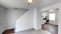 This two-story residence offers a straightforward and functional layout designed for comfortable everyday living. The main level features an open-concept design that connects the living area to the kitchen, creating a cohesive flow between spaces. The kitchen is well-appointed with white cabinetry, stainless steel appliances including a range and oven, and a clean countertop workspace framed by a ventilation hood above the stove. The cabinetry extends to the upper walls, maximizing storage efficiency.

The living area is finished with plush carpeting, providing a warm and comfortable underfoot feel, while the kitchen and adjacent transitional spaces feature hardwood-style flooring that adds a touch of contrast and visual interest. Crisp white walls throughout the main level lend a bright, airy ambiance and serve as a neutral backdrop adaptable to various interior styles.

A staircase with a clean metal handrail leads to the second floor, with the underside of the stairs neatly enclosed to maintain a tidy aesthetic. Ceiling-mounted lighting and well-placed ventilation fixtures contribute to the home's practical design. The overall interior presents a move-in ready environment with a clean, minimalist character suited for a variety of occupants.