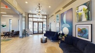 This contemporary lobby serves as a welcoming communal hub, thoughtfully designed to balance elegance with functionality. The space is anchored by soaring ceilings and a striking sputnik-style chandelier with exposed bulbs, casting warm light across the expansive light-toned tile flooring below. Floor-to-ceiling glass transom doors and large flanking windows flood the interior with abundant natural light while providing views to an outdoor balcony and the surrounding community beyond.

The main lounge area is furnished with plush navy blue curved sofas and coordinating accent chairs, arranged around cylindrical drum coffee tables with metallic finishes. A curated gallery wall featuring bold, abstract artwork in shades of blue and teal serves as a dramatic focal point, complemented by fresh orchid arrangements and a large tropical indoor plant that adds a touch of organic warmth.

Adjacent to the main lobby, a private meeting or dining room is accessible via a sliding frosted-glass barn door, equipped with a dining table and chairs for resident gatherings. A secondary entrance area features additional seating and a reception-style workspace with modern lighting. An elevator provides convenient vertical access. Throughout, the design palette of navy, gold, and soft neutrals creates a cohesive, upscale ambiance.