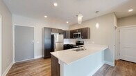 This single-floor residence offers a thoughtfully designed open-concept layout that seamlessly connects the kitchen and living area. The kitchen serves as the centerpiece of the space, featuring a large peninsula with a crisp white countertop, an undermount sink, and dark espresso-toned cabinetry that provides strong visual contrast. A full suite of stainless steel appliances — including a refrigerator, electric range, built-in microwave, and dishwasher — equips the kitchen for everyday functionality. Pendant lighting above the peninsula adds a polished, modern touch alongside recessed ceiling lights throughout.

Wood-look flooring extends through the main living areas and hallways, contributing to a warm, cohesive aesthetic. The living area receives natural light through a glass rear door, creating an inviting atmosphere. A hallway leads to the bedroom, which features neutral-toned carpet, and a dedicated in-unit laundry room housing a full-size front-loading washer and dryer.

Two full bathrooms serve the unit. The primary bathroom includes a double-sink vanity with a wide mirror, a soaking tub, and access to a walk-in closet with built-in shelving. The second bathroom offers a tub-shower combination with clean, white finishes and wood-look flooring. Neutral wall colors and clean architectural lines give the entire space a fresh, contemporary feel.