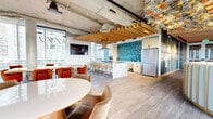 Located at 809 S Washington St in Seattle's Pioneer Square neighborhood, this 2,260-square-foot single-floor amenity space delivers a sophisticated, design-forward environment defined by high exposed ceilings with industrial ductwork, warm wood-tone flooring, and abundant natural light from floor-to-ceiling windows. The open-plan interior is organized into distinct zones that flow seamlessly into one another. A lounge area anchors one end of the space, featuring neutral sofas, a double-sided linear fireplace, and a wall-mounted display screen. Adjacent to it, a dining area with bold red accent chairs and a teal-tiled kitchen bar provides a social gathering point beneath a striking sculptural chandelier of clustered glass globes. A carpeted lounge zone offers intimate seating clusters framed by dramatic large-scale art glass panels etched with abstract forest imagery, complemented by slatted wood ceiling elements. A reception area with a curved blue-and-gold desk serves as a welcoming focal point near the entrance. The space extends outdoors to a wraparound terrace furnished with yellow woven lounge chairs, colorful iridescent glass balustrades, and open views of the Seattle skyline and surrounding tree canopy, creating a seamless indoor-outdoor experience.