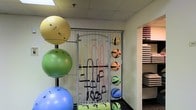 This single-floor fitness space is designed to support a range of workout activities, with an organized and functional layout that makes efficient use of the available area. A central wall-mounted equipment rack serves as the focal point of the room, neatly storing an assortment of color-coded resistance bands with handles, keeping them easily accessible for users. Flanking the rack is a vertical stand holding multiple stability balls in varying sizes and colors, including yellow, green, and blue, offering options for balance and core training exercises. To the right of the rack, a dedicated shelving unit displays a collection of medicine balls in multiple weights, identifiable by their distinct color coding, alongside a neat stack of exercise mats stored on a separate shelf. The ceiling features a drop-tile design, contributing to the room's practical, no-frills aesthetic. The space is finished with dark flooring that complements the utilitarian character of the room. Overall, the layout prioritizes accessibility and organization, making it a well-equipped environment suited for strength, flexibility, and functional fitness training.