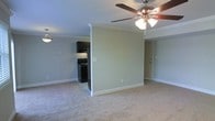 This single-floor residence offers a clean, functional layout designed for comfortable everyday living. The main living area is finished with plush wall-to-wall carpeting in a warm neutral tone, creating a soft and inviting atmosphere. Crown molding runs along the ceiling perimeter, adding a refined architectural detail to the space. A ceiling fan with an integrated light fixture is centrally positioned, providing both ventilation and ambient lighting to the room.

The living area flows naturally into the kitchen, which is partially separated by a half-wall partition that defines each space while maintaining an open, connected feel. The kitchen features dark cabinetry that provides a striking contrast against the light green-gray walls, and is equipped with black appliances including a range and overhead cabinetry. Tile flooring in the kitchen differentiates it from the carpeted living zone.

The walls throughout are painted in a soft, muted sage-green hue, lending a calm and neutral backdrop that suits a variety of furnishing styles. White baseboards and trim provide crisp contrast throughout. A white door is visible near the kitchen area, indicating access to additional rooms or an entryway. Overall, the space presents a tidy, move-in-ready environment with practical design choices and consistent finishes throughout.