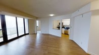 This single-floor residence offers a clean, modern layout designed for comfortable everyday living. The main living area is spacious and open, showcasing warm hardwood-style flooring that extends throughout and crisp white walls that enhance the sense of brightness and airiness. Large windows and a sliding glass door flood the space with natural light, creating a welcoming atmosphere.

A pass-through window connects the living area to the kitchen, maintaining an open feel while defining each space. The kitchen is well-appointed with white cabinetry, dark countertops, and stainless steel appliances, including a microwave, oven range, and dishwasher. Light-toned flooring in the kitchen complements the overall design palette.

A hallway visible from the main living space leads to additional rooms, suggesting a thoughtful and functional floor plan. The neutral color scheme throughout — white walls paired with warm flooring tones — provides a versatile backdrop suited to a variety of interior styles. The overall ambiance is clean, contemporary, and move-in ready, making it an appealing and practical living space.