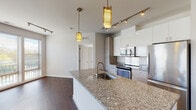 This single-floor residence showcases a contemporary open-concept design that seamlessly connects the kitchen and living areas. The kitchen serves as a central hub, featuring a large granite-topped island with an undermount sink, stainless steel appliances including a refrigerator, range, microwave, and dishwasher, and a combination of white upper cabinets and light wood lower cabinetry. Stylish pendant lights with gold accents hang above the island, complemented by track lighting overhead. Rich dark hardwood flooring extends throughout the main living and kitchen areas, adding warmth to the modern aesthetic.

The living area is bright and open, benefiting from an expansive bank of floor-to-ceiling windows fitted with horizontal blinds that offer outdoor views and generous natural light. The bedrooms feature soft carpeting, neutral-toned walls, and windows with outdoor views, creating comfortable, light-filled retreats. Two bathrooms serve the unit: one includes a soaking tub with subway tile surround and a dark wood vanity with granite countertop, while the second features a glass-enclosed walk-in shower and built-in shelving. A stacked in-unit washer and dryer adds everyday convenience to this well-appointed space.