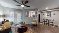 This single-floor residence offers a cohesive and modern living experience with warm neutral tones and durable wood-look flooring that flows seamlessly throughout the space. The open-concept living area serves as the heart of the home, featuring a ceiling fan overhead, large French doors that invite abundant natural light, and a cozy seating arrangement anchored by a tufted leather ottoman. A decorative wall display and an assortment of mirrors add character and depth to the space. Adjacent to the living area, a dining nook provides a dedicated space for meals, while the pass-through kitchen connects effortlessly to the main living zone. The kitchen is outfitted with crisp white cabinetry, white countertops, a stainless steel sink with a matte black faucet, a stainless steel dishwasher, and a gas range, all complemented by subway tile backsplash and ample natural light from a window above the counter. The bedroom is a calm and restful retreat, featuring its own ceiling fan, wood-look flooring, and a window with blinds for light control. The bathroom includes a full tub-shower combination with decorative curtains, a toilet, white cabinetry with overhead storage, and wood-look flooring, maintaining the consistent aesthetic found throughout the home.