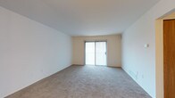 This single-floor space features a spacious, open living area characterized by its clean, minimalist design. The room is finished with wall-to-wall neutral-toned carpeting that adds warmth underfoot while contributing to a quiet, comfortable atmosphere. The walls are painted in a soft, off-white tone that enhances the sense of openness and pairs well with the light-colored ceiling.

A prominent sliding glass door serves as the focal point of the main living area, fitted with vertical blinds that allow for flexible light control. The door provides access to the outdoors and introduces an abundance of natural light, brightening the interior throughout the day. Baseboard vents are positioned along the walls, indicating a forced-air climate control system for year-round comfort.

The layout is straightforward and versatile, offering an open canvas suitable for a variety of furniture arrangements. The absence of clutter or excessive architectural detailing gives the space a practical, functional character. Overall, the space presents a tidy, well-maintained environment with neutral finishes that appeal to a wide range of tastes and uses.