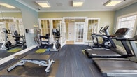 This single-floor amenity facility at 3502 Jenks Ave in Panama City, Florida, offers a well-appointed fitness center and business lounge designed to serve the needs of its users. The layout is thoughtfully organized, with a light-tiled corridor connecting the various spaces, accented by sage green walls, crown molding, and white trim throughout.

The fitness center is the centerpiece of the facility, featuring a generous selection of cardio and strength equipment, including treadmills, elliptical machines, a stationary bike, a multi-station cable machine, free weights, and a weight rack. The room is finished with commercial-grade carpet tile flooring and benefits from abundant natural light streaming through multiple windows with exterior views. A ceiling fan, wall-mounted television, and decorative crown molding contribute to a comfortable workout environment. French doors with glass panels connect the fitness space to the corridor, enhancing openness and flow.

Adjacent to the fitness area is a cozy business lounge, furnished with a built-in desk, computer workstation, printer, and a cushioned bench seat — ideal for remote work or study. Tasteful artwork and neutral-toned carpeting create a calm, focused atmosphere. The facility also includes dual stainless steel drinking fountains set into a tiled niche, providing a practical and accessible convenience for all users.