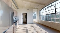 Located in the heart of Downtown Los Angeles, this single-floor industrial loft at 438 S San Pedro St blends historic character with modern updates. The space showcases its warehouse roots through exposed ductwork and piping along the high ceilings, polished and raw concrete flooring throughout, and striking steel-framed windows — including signature arched varieties — that flood the interior with abundant natural light. Sunlight casts geometric shadow patterns across the floors, adding a dynamic visual element to the open layout.

The main living area is spacious and open-concept, flowing naturally into a contemporary kitchen equipped with wood-toned cabinetry, dark stone countertops, a tile backsplash, and stainless steel appliances including a refrigerator, range, microwave, and dishwasher. A long corridor lined with floor-to-ceiling steel-framed windows and warm wood-framed translucent sliding doors connects the various areas of the space, offering continuous city views of the surrounding Downtown LA skyline. Additional rooms provide flexible use as bedrooms, offices, or creative studios, each benefiting from the same bright, airy ambiance. The combination of raw industrial elements and clean white walls gives this loft a versatile, gallery-like quality throughout.