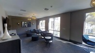 This contemporary lounge space is thoughtfully designed to offer a comfortable and inviting atmosphere. The single-floor layout features a well-appointed seating area furnished with modern barrel-style armchairs and rounded ottomans arranged in casual groupings, encouraging relaxed conversation and social interaction. Accent tables, including sleek bistro-style and marble-top round tables, are strategically placed throughout to provide functional surfaces without interrupting the open flow of the space. The flooring consists of patterned carpet in deep charcoal tones, complementing the neutral taupe and gray wall palette and lending a cohesive, sophisticated aesthetic. Overhead, globe pendant chandeliers with a warm metallic finish serve as elegant focal points, casting a warm ambient glow that contrasts beautifully with the abundant natural light streaming in through the large floor-to-ceiling windows along the exterior wall. These expansive windows offer views of an adjacent outdoor balcony and courtyard area, creating a seamless visual connection to the outside. A framed piece of wall art adds a touch of artistic character to the space. Overall, the lounge strikes a balance between modern design and approachable comfort, making it an ideal gathering and relaxation area.