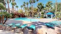 This expansive resort-style pool area offers a striking combination of natural and designed elements that create a true oasis atmosphere. The centerpiece is a generously sized, freeform swimming pool with crystal-clear turquoise water, framed by a dramatic red rock formation complete with a waterfall feature and a remarkable grotto — a cave-like overhang that extends over a portion of the pool, adding a sense of adventure and seclusion. Tall palm trees and mature shade trees line the perimeter, providing a lush, tropical canopy that filters natural sunlight throughout the space. The surrounding deck is outfitted with numerous teal-colored lounge chairs and patio umbrellas, offering plenty of space for sunbathing and relaxation. A separate, circular spa with bubbling water is nestled nearby, accompanied by a covered pergola seating area — ideal for unwinding in a more intimate setting. The architectural backdrop features a Mediterranean-influenced building with arched windows and stucco finishes, complementing the warm desert landscape. The overall ambiance is vibrant yet relaxing, evoking the feel of a private resort retreat under the clear blue skies of a sunny, arid climate.