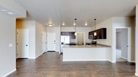 This single-floor space showcases an open-concept layout that connects the living area and kitchen in a seamless, functional design. The kitchen is a standout feature, equipped with rich dark wood cabinetry that contrasts elegantly against light granite countertops. Stainless steel appliances, including a French door refrigerator, microwave, and range, add a modern, polished look. A peninsula with a white base and granite surface extends into the living space, offering additional prep area and casual seating potential. Pendant lighting with dark fixtures hangs above the peninsula, providing both task lighting and a stylish focal point. Recessed lighting throughout the space ensures consistent, even illumination. The flooring is a deep-toned hardwood that runs continuously through both areas, anchoring the open layout with warmth and cohesion. Neutral white walls keep the space feeling bright and versatile. A hallway near the entryway leads to additional areas of the unit, while the solid white entry door features a contemporary design. Overall, the space balances modern finishes with practical functionality.