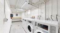 Located at 10740 Blix St in Los Angeles, California, this shared laundry facility is part of the Cape Cod Garden community. The room is constructed with white-painted concrete block walls and finished with durable light-toned flooring, giving the space a clean and utilitarian aesthetic throughout.

The facility is equipped with a row of commercial-grade Speed Queen front-loading washers and matching dryers, arranged along one wall to maximize efficiency and ease of use. Exposed white-painted plumbing and ventilation pipes run along the ceiling, which is illuminated by fluorescent lighting fixtures that ensure bright, even coverage across the entire room. A wall-mounted paper towel dispenser and a fire extinguisher are also present, contributing to the room's practical, safety-conscious design.

At the far end of the room, a built-in wooden folding shelf provides a dedicated surface for sorting and folding laundry. Directly above it, a clearly posted "Cape Cod Garden Laundry Rules" sign outlines guidelines for residents' use of the facility. A louvered door provides access to the room, adding a functional architectural detail to the otherwise straightforward space. Floor drains are also visible, facilitating easy maintenance and cleaning of the facility.