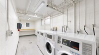 Located at 10740 Blix St in Los Angeles, California, this shared laundry facility is part of the Cape Cod Garden community. The room is constructed with white-painted concrete block walls and finished with durable light-toned flooring, giving the space a clean and utilitarian aesthetic throughout.

The facility is equipped with a row of commercial-grade Speed Queen front-loading washers and matching dryers, arranged along one wall to maximize efficiency and ease of use. Exposed white-painted plumbing and ventilation pipes run along the ceiling, which is illuminated by fluorescent lighting fixtures that ensure bright, even coverage across the entire room. A wall-mounted paper towel dispenser and a fire extinguisher are also present, contributing to the room's practical, safety-conscious design.

At the far end of the room, a built-in wooden folding shelf provides a dedicated surface for sorting and folding laundry. Directly above it, a clearly posted "Cape Cod Garden Laundry Rules" sign outlines guidelines for residents' use of the facility. A louvered door provides access to the room, adding a functional architectural detail to the otherwise straightforward space. Floor drains are also visible, facilitating easy maintenance and cleaning of the facility.