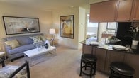This single-floor residence showcases an open-concept design that integrates the living, dining, and kitchen spaces into one cohesive, functional layout. The living area is carpeted and comfortably sized, anchored by a contemporary sofa adorned with decorative pillows and paired with a sleek glass-and-chrome coffee table. Framed artwork on the warm-toned walls adds visual interest and a curated aesthetic throughout.

A dark-finished kitchen peninsula with a granite or glass countertop serves as a natural divider between the kitchen and living areas, accompanied by a stylish black upholstered bar stool. The peninsula provides both additional prep space and a casual dining option. Dark wood cabinetry is visible in the kitchen, complementing the countertop tones.

Neutral, light-colored carpet flows throughout the main living space, contributing to a warm and inviting atmosphere. A window with a roman shade allows natural light to filter into the area. Through an open passageway, a bedroom is partially visible, featuring a neatly made bed and access to what appears to be a vanity or bathroom area, suggesting a practical and well-organized floor plan throughout the residence.