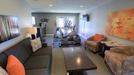 This single-floor amenity space serves a dual purpose, thoughtfully combining a fitness area with a relaxed lounge environment. The room is finished with neutral gray walls, recessed ceiling lighting, and a textured ceiling, creating a clean and welcoming atmosphere throughout.

The fitness portion of the space is equipped with multiple cardio machines, including ellipticals and a stationary bike, all positioned on a protective dark floor mat near a set of windows that allow natural light to filter in. The natural light enhances the energy of the workout area while keeping the space feeling open and airy.

Transitioning seamlessly into the lounge area, the space features a dark wood coffee table, a plush gray sofa, and a patterned armchair with orange accent pillows, adding a pop of color and warmth. A floor lamp provides soft ambient lighting, complementing the recessed overhead fixtures. A wall-mounted flat-screen TV is positioned for easy viewing from the seating area, making it a comfortable spot to unwind.

A small refreshment or coffee station is tucked neatly along one wall, offering added convenience for residents or guests. The combination of functional fitness equipment and inviting lounge furnishings makes this a versatile, well-appointed communal amenity space.