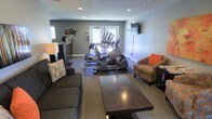 This single-floor amenity space serves a dual purpose, thoughtfully combining a fitness area with a relaxed lounge environment. The room is finished with neutral gray walls, recessed ceiling lighting, and a textured ceiling, creating a clean and welcoming atmosphere throughout.

The fitness portion of the space is equipped with multiple cardio machines, including ellipticals and a stationary bike, all positioned on a protective dark floor mat near a set of windows that allow natural light to filter in. The natural light enhances the energy of the workout area while keeping the space feeling open and airy.

Transitioning seamlessly into the lounge area, the space features a dark wood coffee table, a plush gray sofa, and a patterned armchair with orange accent pillows, adding a pop of color and warmth. A floor lamp provides soft ambient lighting, complementing the recessed overhead fixtures. A wall-mounted flat-screen TV is positioned for easy viewing from the seating area, making it a comfortable spot to unwind.

A small refreshment or coffee station is tucked neatly along one wall, offering added convenience for residents or guests. The combination of functional fitness equipment and inviting lounge furnishings makes this a versatile, well-appointed communal amenity space.