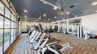 Located at 110 1 Mile Parkway in Nashville, TN, this fitness center offers a spacious, single-floor exercise space designed to support a comprehensive workout experience. The room is generously sized, providing ample floor space for a wide variety of equipment and free movement throughout.

The interior design features a striking dark slate-blue ceiling paired with crisp white walls, creating a modern and energizing atmosphere. Recessed lighting throughout ensures the space is well-lit, while patterned, multi-tone carpet tiles add texture and a contemporary aesthetic underfoot.

A row of Matrix-brand treadmills and elliptical machines lines one side of the room, ideal for cardiovascular training. A rowing machine adds additional cardio variety. On the strength side, the facility includes cable and pulley machines, multi-station weight systems, a full dumbbell rack ranging across multiple weight increments, adjustable benches, and a medicine ball storage rack. Three large framed mirrors are mounted along one wall, enhancing the sense of space and supporting proper form during workouts.

A standout architectural feature is the oversized glass garage-style window wall, which floods the space with natural light and provides views of an outdoor green area. A dedicated entry door with an exterior view further connects the interior to the outdoors, contributing to an open, airy ambiance throughout the facility.