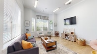 Located at 7221 US Highway 70 S in Nashville, TN, this single-room living space offers a thoughtfully designed and inviting atmosphere. The room features light hardwood flooring and soft, pale gray walls that create a clean, airy backdrop throughout. Crown molding adds a refined architectural detail, while elevated ceilings contribute to a sense of openness beyond the room's compact footprint.

A defining feature is the angled bay window area in the corner, dressed with white horizontal blinds that allow generous natural light to filter in while framing views of the lush, green outdoor surroundings. Beneath the windows, a pair of curved, mid-century modern accent chairs provide a cozy reading or conversation nook.

The entertainment wall is anchored by a wall-mounted flat-screen television positioned above a rustic barn-style media console, which adds warmth and texture to the space. A plush, dark purple mid-century sofa accented with vibrant orange and teal throw pillows serves as the centerpiece seating, paired with a sturdy wooden coffee table atop a vintage-style area rug.

A standout decorative element is the oversized wall-mounted Scrabble board, adding a playful, social dimension to the room. Overhead, linear fluorescent lighting fixtures provide consistent illumination. The space is completed by decorative lettering, framed artwork, and thoughtfully curated accessories that give the room a warm, eclectic personality.