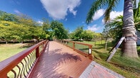 This outdoor space features a well-crafted walkway bridge constructed with rich, warm-toned hardwood decking that extends in a gentle curve through a lush tropical landscape. The bridge is lined with ornate railing systems combining sturdy wooden posts and decorative black metal balusters with a distinctive wave pattern, lending both safety and aesthetic appeal to the structure. Built-in low-profile lighting fixtures are integrated along the railing posts, suggesting the walkway remains functional and visually attractive into the evening hours. The bridge transitions from a herringbone-patterned brick pathway, adding a refined design element at its entry point. Surrounding the structure, the landscape is rich with mature tropical vegetation, including towering palm trees and a dense canopy of broad-leafed trees that provide shade and a vibrant green backdrop. An open grassy area is visible beyond the bridge, bordered by additional trees and a fence line in the distance. The open sky above, dotted with scattered clouds, floods the space with natural light, enhancing the tropical ambiance. Overall, this walkway bridge offers a visually appealing and functional outdoor passage within a well-maintained, verdant setting.