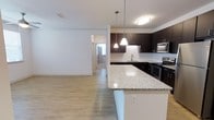 This single-floor, three-bedroom, two-bathroom residence offers a thoughtfully designed open-concept layout that maximizes both functionality and livable space. The kitchen serves as a central highlight, featuring rich dark cabinetry that contrasts elegantly with light speckled granite countertops. A large kitchen island provides additional prep and dining space, while pendant lighting adds a refined, modern touch. Stainless steel appliances—including a gas range, built-in microwave, and refrigerator—complete the well-equipped cooking area. A subway tile backsplash and undermount sink contribute to the kitchen's clean, contemporary aesthetic. Light wood-tone flooring flows throughout the main living areas, creating a warm, cohesive atmosphere. The open connection between the kitchen and living area allows for easy interaction and entertaining. The floor plan, as seen in the 3D model, reveals a well-organized layout with three distinct bedrooms and two full bathrooms, offering comfortable accommodations for residents. The bright, neutral walls and ample lighting throughout contribute to an overall airy and welcoming ambiance.