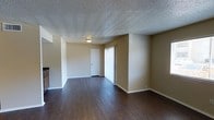 This single-floor space offers a clean and functional layout designed for comfortable everyday living. The main living area is defined by rich, dark hardwood-style flooring that extends throughout, providing a warm contrast against the neutral beige walls and crisp white trim and baseboards. The textured popcorn ceiling adds a classic finishing touch overhead, while a flush-mount ceiling light provides central illumination to the room.

The open living area is notably spacious, allowing for a variety of furniture arrangements. A sliding glass door fitted with vertical blinds lines one wall, offering access to the outdoors and inviting natural light into the interior. Adjacent to the sliding door, a solid white interior door leads to an additional room or closet space, contributing to the overall functionality of the unit.

A partial view of cabinetry visible at the edge of the frame hints at a nearby kitchen area, suggesting a practical and connected floor plan. The walls are finished in a soft, warm tan tone that complements the flooring and creates a cohesive, understated aesthetic throughout. Overall, the space presents a straightforward, well-maintained interior with durable finishes and a layout that balances openness with defined living zones.