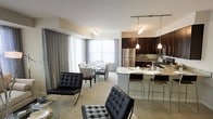 This single-floor residence showcases a thoughtfully designed open-concept layout that seamlessly connects the living, dining, and kitchen areas into one cohesive and functional space. The living area is anchored by sleek, tufted black leather Barcelona-style chairs that lend a contemporary and sophisticated character to the room. Large windows dressed with floor-to-ceiling grey curtains flood the interior with natural light, creating a bright and airy ambiance throughout. The dining area accommodates a glass-top table surrounded by upholstered chairs, making it an elegant setting for gatherings. The modern kitchen is a standout feature, equipped with rich dark espresso cabinetry, stainless steel appliances, and a granite-topped island with bar seating for casual dining. Pendant lighting above the island and track lighting overhead provide both functional illumination and a refined aesthetic touch. The neutral tones of the walls and flooring unify the space, offering a clean, contemporary backdrop that complements the bold furnishings. The overall ambiance is polished and inviting, balancing modern design elements with a warm, livable atmosphere suited for both everyday living and entertaining.