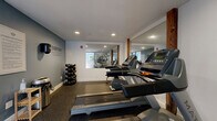 Located at 219 E Bank St in Petersburg, Virginia, this fitness center is a well-appointed exercise room designed to provide a focused and functional workout environment. The space features durable rubber flooring throughout, offering both safety and comfort during exercise. A row of Matrix treadmills serves as the centerpiece for cardio training, positioned to take advantage of the room's layout and natural light filtering through frosted floor-to-ceiling glass entry doors.

A full dumbbell rack lines one wall, offering a range of free weights for strength training, accompanied by an adjustable workout bench for versatile exercises. A large wall-mounted mirror visually expands the space and allows users to monitor their form. The mirror is framed by exposed wooden posts, adding a warm, industrial accent to the otherwise modern interior.

The walls are painted in a calming slate blue and white palette, with a bold motivational phrase — "Everyday is a Good Day to Workout" — displayed prominently, enhancing the energizing atmosphere. Recessed ceiling lighting ensures the room is well-lit at all times. A sanitation station with paper towels and a waste receptacle reflects attention to cleanliness. A posted fitness center rules board near the entrance rounds out this practical and inviting exercise facility.