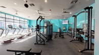 This modern fitness center is designed to support a comprehensive range of workout routines across multiple distinct zones. The main gym floor is defined by vibrant teal accent walls, rubber flooring, and a wall of floor-to-ceiling windows that flood the space with natural light and street views. It is outfitted with a full row of treadmills and ellipticals along the window line, a multi-functional cable machine, adjustable benches, and a well-stocked dumbbell rack backed by full-length mirrors. Mounted flat-screen TVs and multiple ceiling fans contribute to a comfortable, modern workout environment. A separate spin studio features wood-look flooring, a row of stationary cycling bikes, and an energizing typographic mural covering an entire wall with fitness-themed words in bold, colorful lettering. An additional studio room with wood-look flooring, barre rails, large mirrors, a wall-mounted TV, and stability balls supports group fitness, stretching, and barre classes. A dedicated locker area with built-in lockers, rubber flooring, and ceiling fans provides convenient storage. Throughout the facility, the consistent teal color palette and recessed lighting create a cohesive, energetic ambiance suited for all fitness levels.