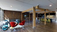 This single-floor space showcases a compelling blend of industrial and contemporary design elements. Exposed brick walls in rich, warm tones serve as a defining architectural feature throughout, complemented by natural wood beam columns that add structural character and visual warmth. The dark hardwood flooring grounds the space and ties together its distinct functional zones.

The lounge area is furnished with a grey sofa and striking red swan-style accent chairs arranged around a low white coffee table, creating an inviting and stylish seating vignette. Adjacent to this, a high-top round table with bar stools provides a more casual standing or perching option, ideal for informal gatherings or quick meetings.

Further into the space, a long communal bar-height table is lined with bright yellow industrial stools, adding a bold pop of color against the darker tones of the brick and flooring. A wall-mounted flat-screen TV is positioned for visibility across the space, suggesting a media or presentation function. Pendant lighting and recessed fixtures illuminate the area, while a white drop ceiling contrasts with the raw brick to balance the industrial aesthetic with a clean, modern finish.