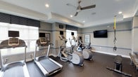 This fitness center offers a clean, well-appointed space designed to support an active lifestyle. The room is finished with a neutral gray and blue color scheme, accented by a decorative horizontal stripe that runs along the perimeter walls, lending the space a polished, purposeful aesthetic. Recessed lighting and a centrally mounted ceiling fan with a brushed nickel finish work together to maintain a bright and comfortable atmosphere during workouts. Large windows along the front wall invite generous natural light while providing views of the outdoors, keeping the environment open and energizing. The space is equipped with a lineup of cardio machines, including treadmills and elliptical trainers, each featuring individual digital display screens for tracking workout metrics. A wall-mounted flat-screen television is positioned for easy viewing from any piece of equipment. The floors are covered in durable dark rubber flooring, well-suited for high-traffic fitness use. Roman shades in a coordinating patterned fabric dress the windows, adding a refined touch to the utilitarian space. A framed piece of wall art contributes to the overall welcoming ambiance. This fitness center balances functionality and design thoughtfully, creating a motivating environment for daily exercise.
