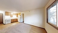 This single-floor unit features an open-concept layout that seamlessly connects the living area to the kitchen. The living space is finished with wall-to-wall neutral carpet, providing a soft underfoot feel, while cream-colored walls and flush ceiling lighting create a bright, clean atmosphere. Multiple electrical outlets are positioned along the walls, offering practical flexibility for furniture arrangement.

The kitchen is situated in the corner and is defined by warm honey-toned wood cabinetry that provides ample storage. It is equipped with a white refrigerator and features a sink set beneath under-cabinet lighting, which illuminates the workspace efficiently. The kitchen floor transitions to smooth hard-surface flooring, contrasting with the carpeted living area. A doorway within the kitchen leads further into the unit, suggesting additional rooms beyond what is visible.

Overall, the space presents a straightforward, functional design with a neutral color palette throughout. The open sightlines between the living and kitchen areas contribute to an efficient use of space, making it well-suited for everyday living.