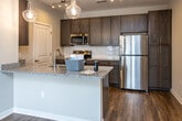 Located at 100 Vintage Alcoa Way in Alcoa, Tennessee, this single-story residence offers a thoughtfully designed open-concept layout with consistent wood-look flooring throughout the main living areas. The spacious living room is filled with natural light from large windows dressed with blinds and is equipped with a ceiling fan, creating a comfortable and airy atmosphere. A built-in entryway bench with wall hooks provides a practical transition space near the front door.

The kitchen is a standout feature, showcasing rich dark-toned cabinetry, granite countertops, a subway tile backsplash, and a full suite of stainless steel appliances including a refrigerator, electric range, and microwave. Track lighting and elegant pendant lights illuminate the space, while the peninsula with a granite overhang connects the kitchen seamlessly to the living and dining areas.

The bedroom continues the warm flooring theme and includes a ceiling fan and a window with natural light and exterior views. The bathroom is clean and functional, featuring tile flooring, a walk-in shower with a curved rod, granite vanity countertop, and a well-organized linen closet with wire shelving. The unit presents a modern, neutral palette throughout, offering a move-in ready space with practical finishes and efficient use of layout.